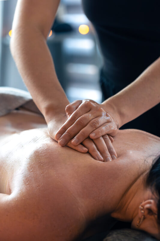 Stacked hands press gently into the center of the back with oil. Warm spa mood turns wellness into a stylish daily habit.